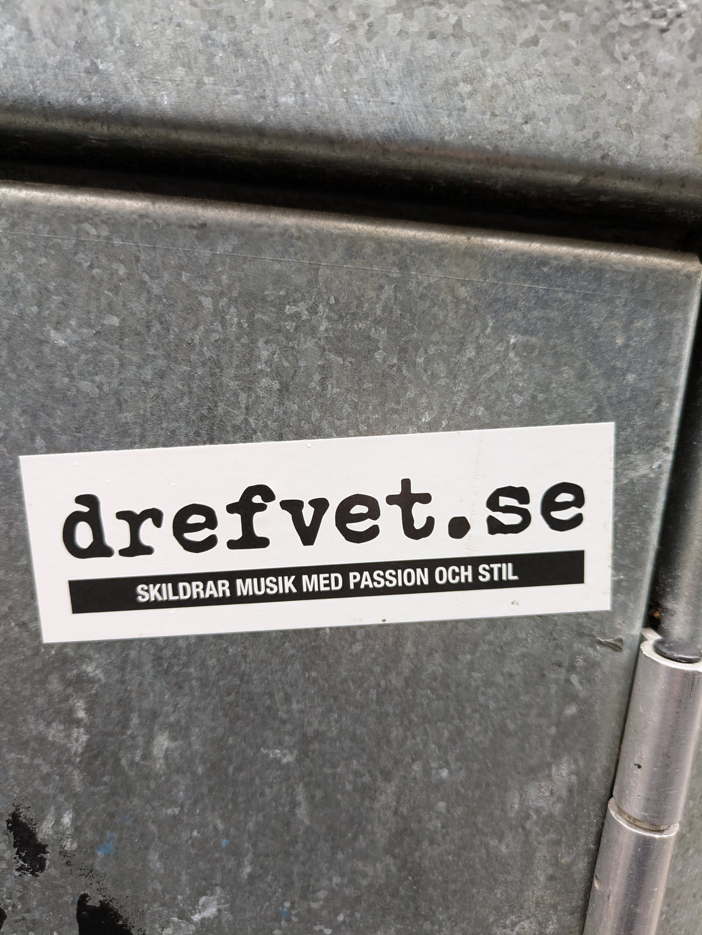 A white sticker with black text on a gray metal surface. The text on the sticker says "dreftvet.se" in a large font and "Skildrar musik med passion och stil" in a smaller font below.