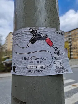 A sticker on a pole advertising a tattoo parlor called Bang Em Out Tattoo's.  The sticker features a cartoon illustration of a tattoo gun and a small character.
