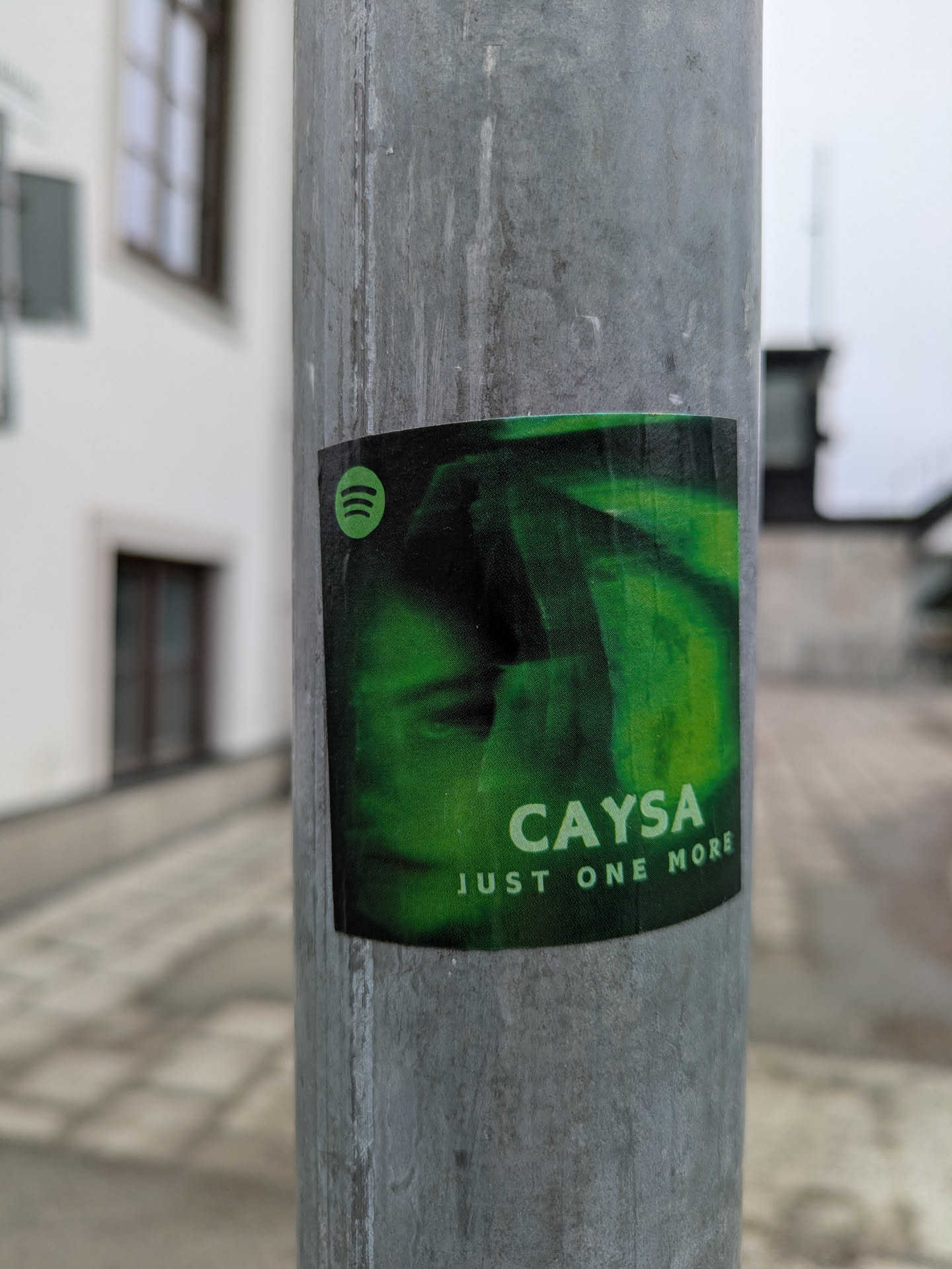 A rectangular sticker with rounded corners is affixed to a light pole. The sticker features the Spotify logo in the upper left corner, indicating the artist CaySA is available on the platform. The sticker's design is predominantly green, featuring a stylized image of the artist's face. Below the artist's name, the text 'JUST ONE MORE' is printed. The light pole is gray, and the background is out of focus, showing a blurred urban or suburban environment.
