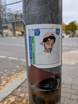 A rectangular sticker designed like a UEFA Champions League Panini collectible card adhered to a metal street pole. It features a portrait of a young man looking downward while holding a cigarette or toothpick between his lips, wearing a pink backwards baseball cap and a light-colored sweatshirt. The card frame includes the Champions League trophy and starball logos at the top, a QR code in the upper-left, and a side banner reading 'PLAYER TO WATCH' above 'OCEANB'.