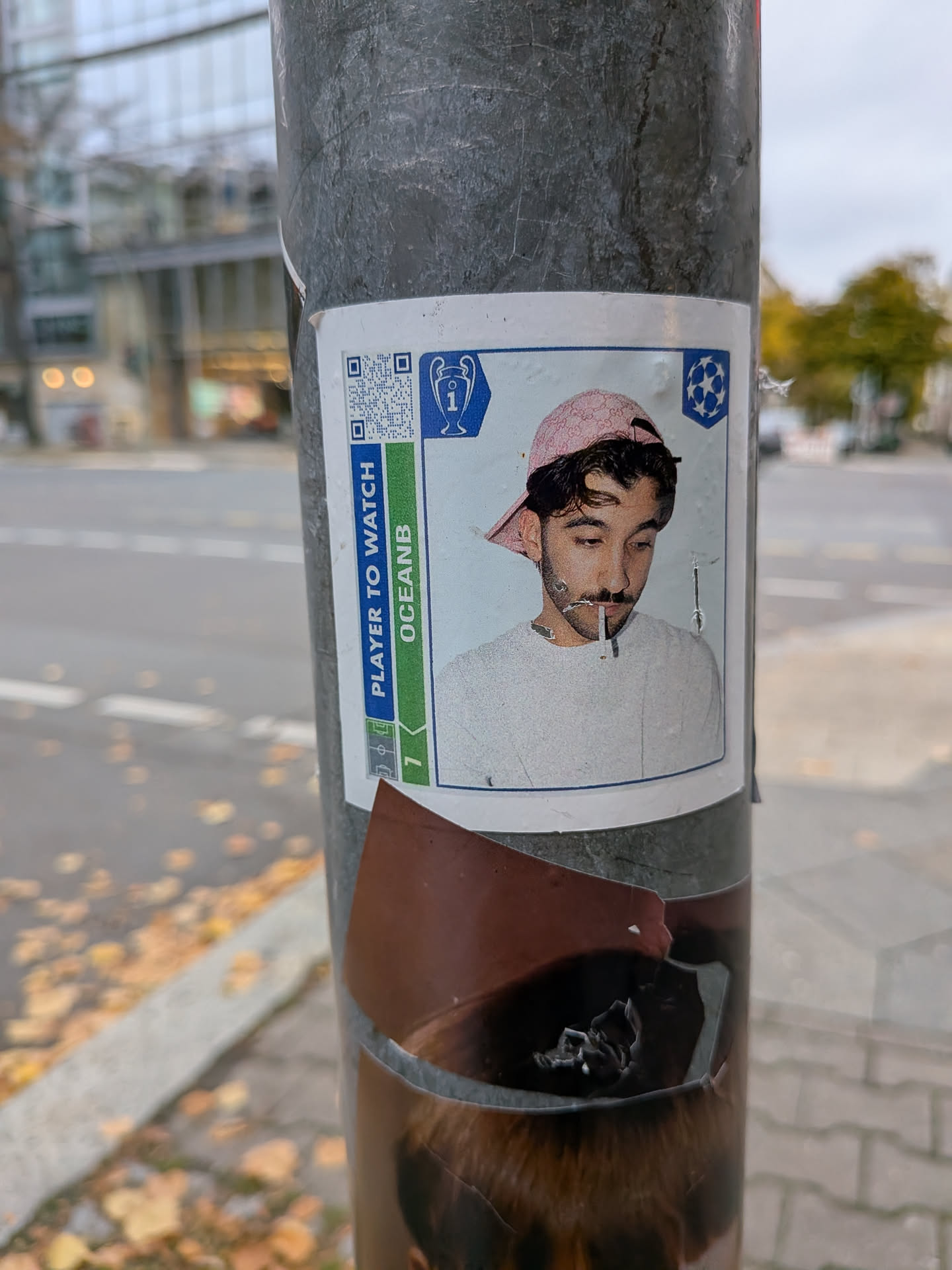 A rectangular sticker designed like a UEFA Champions League Panini collectible card adhered to a metal street pole. It features a portrait of a young man looking downward while holding a cigarette or toothpick between his lips, wearing a pink backwards baseball cap and a light-colored sweatshirt. The card frame includes the Champions League trophy and starball logos at the top, a QR code in the upper-left, and a side banner reading 'PLAYER TO WATCH' above 'OCEANB'.