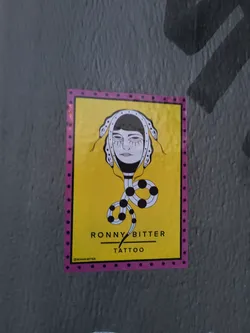 Yellow and pink sticker with illustration of a woman's face with snakes for hair. The text "Ronny Bitter Tattoo" is at the bottom of the sticker.
