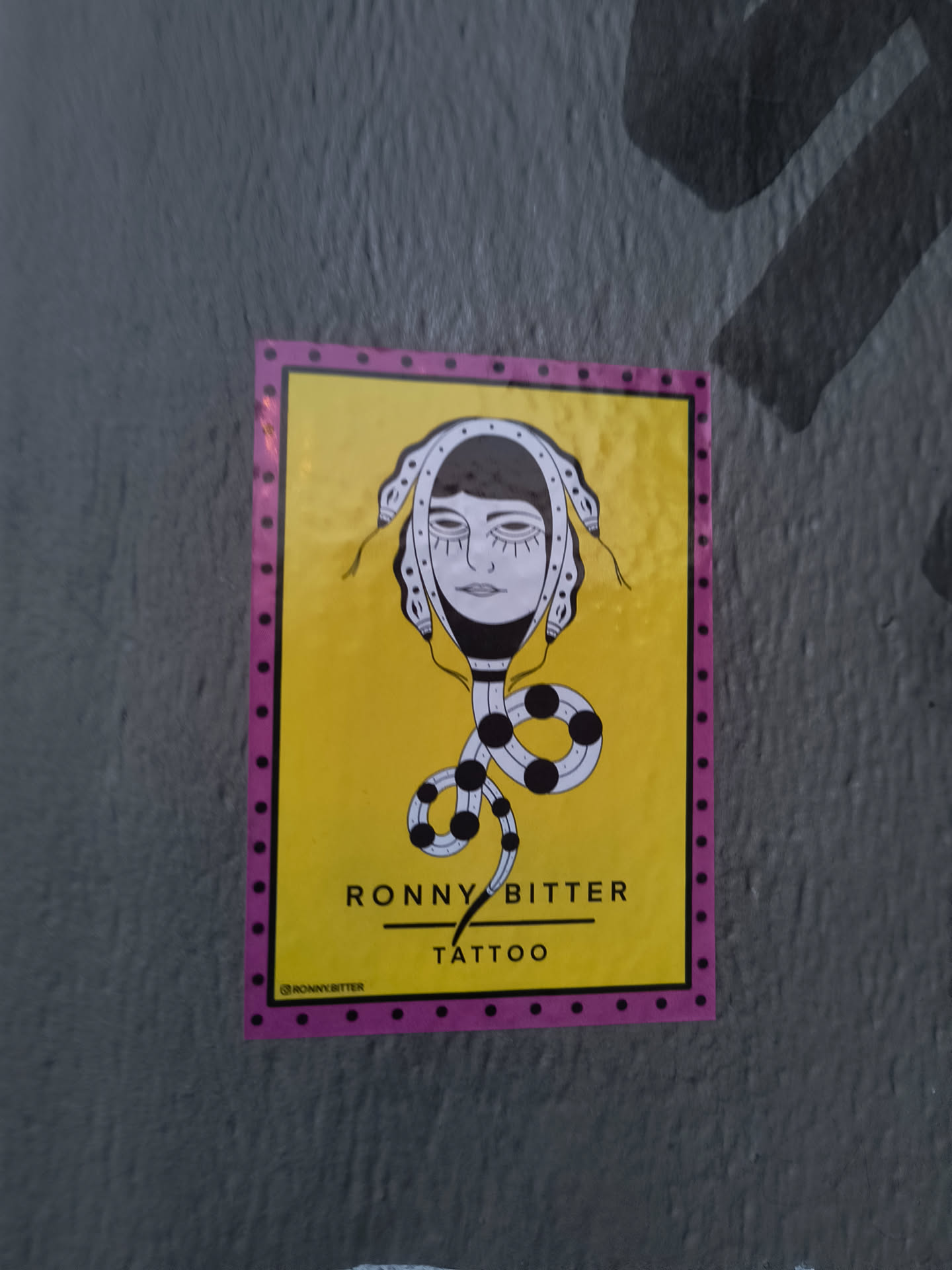 Yellow and pink sticker with illustration of a woman's face with snakes for hair. The text "Ronny Bitter Tattoo" is at the bottom of the sticker.