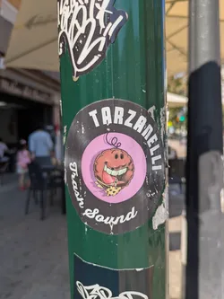 The sticker is round with a black background and a pink center. It shows a cartoon character with a large smile eating a slice of pizza. Text around the circle reads "Tarzanelli Trash Sound".