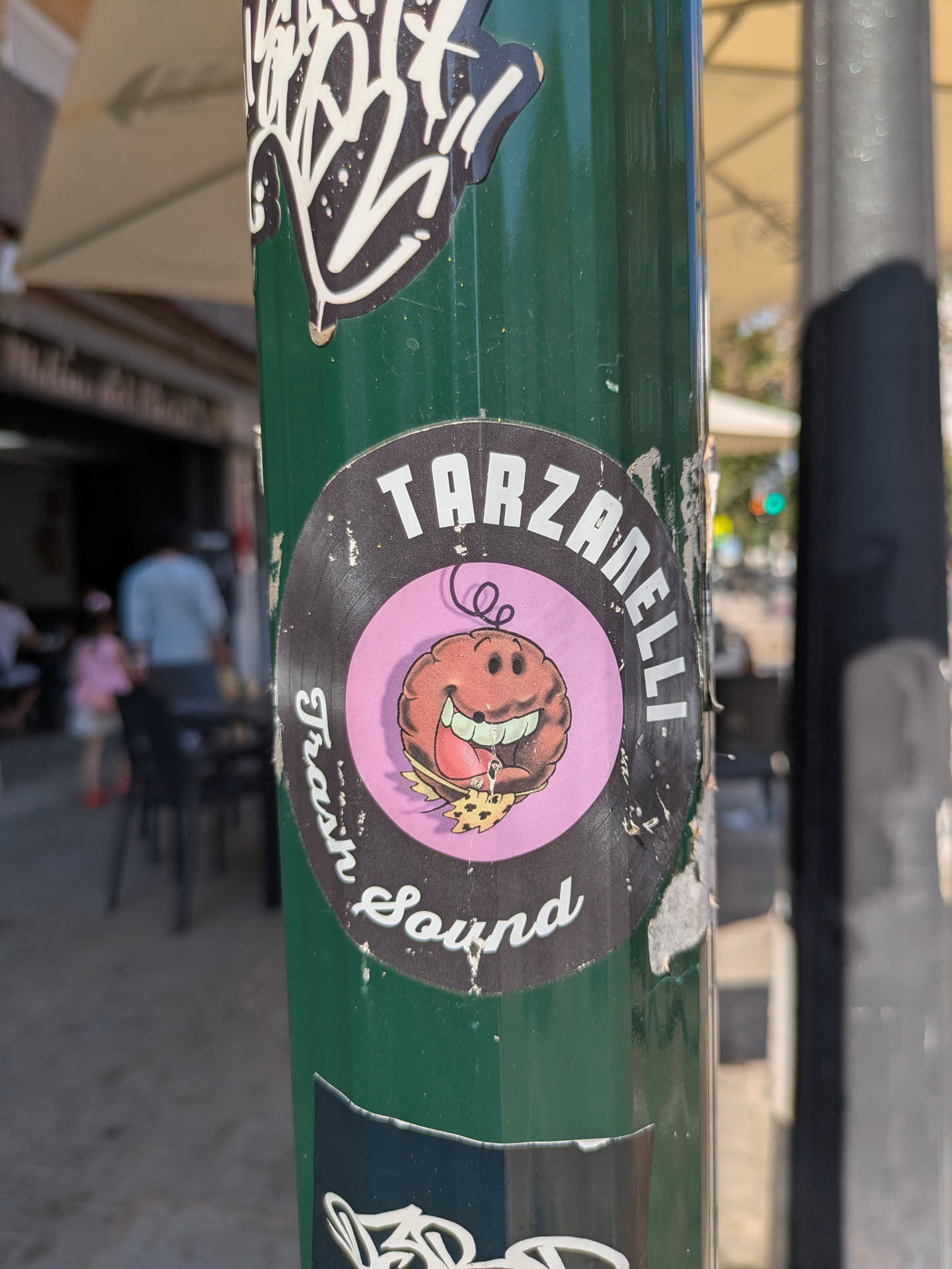 The sticker is round with a black background and a pink center. It shows a cartoon character with a large smile eating a slice of pizza. Text around the circle reads "Tarzanelli Trash Sound".