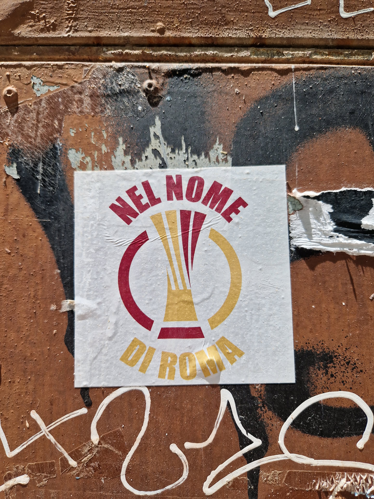 A sticker featuring a logo with the text "Nel Nome Di Roma" (In the Name of Rome) and a graphic that seems to depict a trophy or cup. The sticker is affixed to a weathered, graffiti-covered surface.