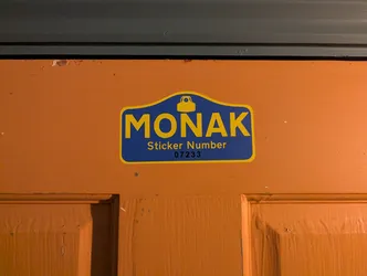 Street sticker A shaped sticker designed in the style of a traditional Parisian street name plaque. It features a rich royal blue background with a thin yellow border. At the top arch is a stylized yellow spray can cap nozzle. Beneath it, the word 'MONAK' is printed in large bold yellow sans-serif capitals. Underneath reads 'Sticker Number' in yellow, followed by stamped black numbering '07233'. The sticker is applied to a bright orange painted wooden door.