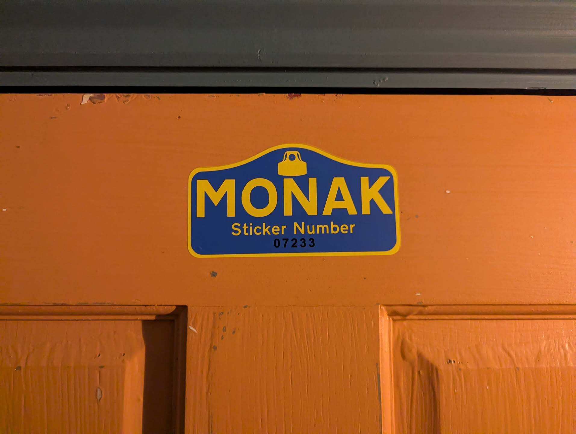 A shaped sticker designed in the style of a traditional Parisian street name plaque. It features a rich royal blue background with a thin yellow border. At the top arch is a stylized yellow spray can cap nozzle. Beneath it, the word 'MONAK' is printed in large bold yellow sans-serif capitals. Underneath reads 'Sticker Number' in yellow, followed by stamped black numbering '07233'. The sticker is applied to a bright orange painted wooden door.