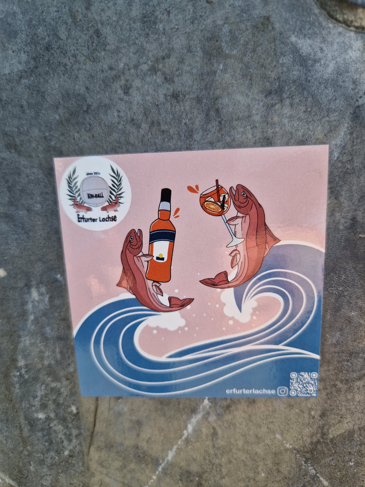 A sticker featuring two salmon toasting with an Aperol Spritz in the middle of an ocean wave. It has a minimalist design with a pink background and blue waves. There is a small logo in the top left corner. The sticker also includes a QR code linking to the artist's Instagram.