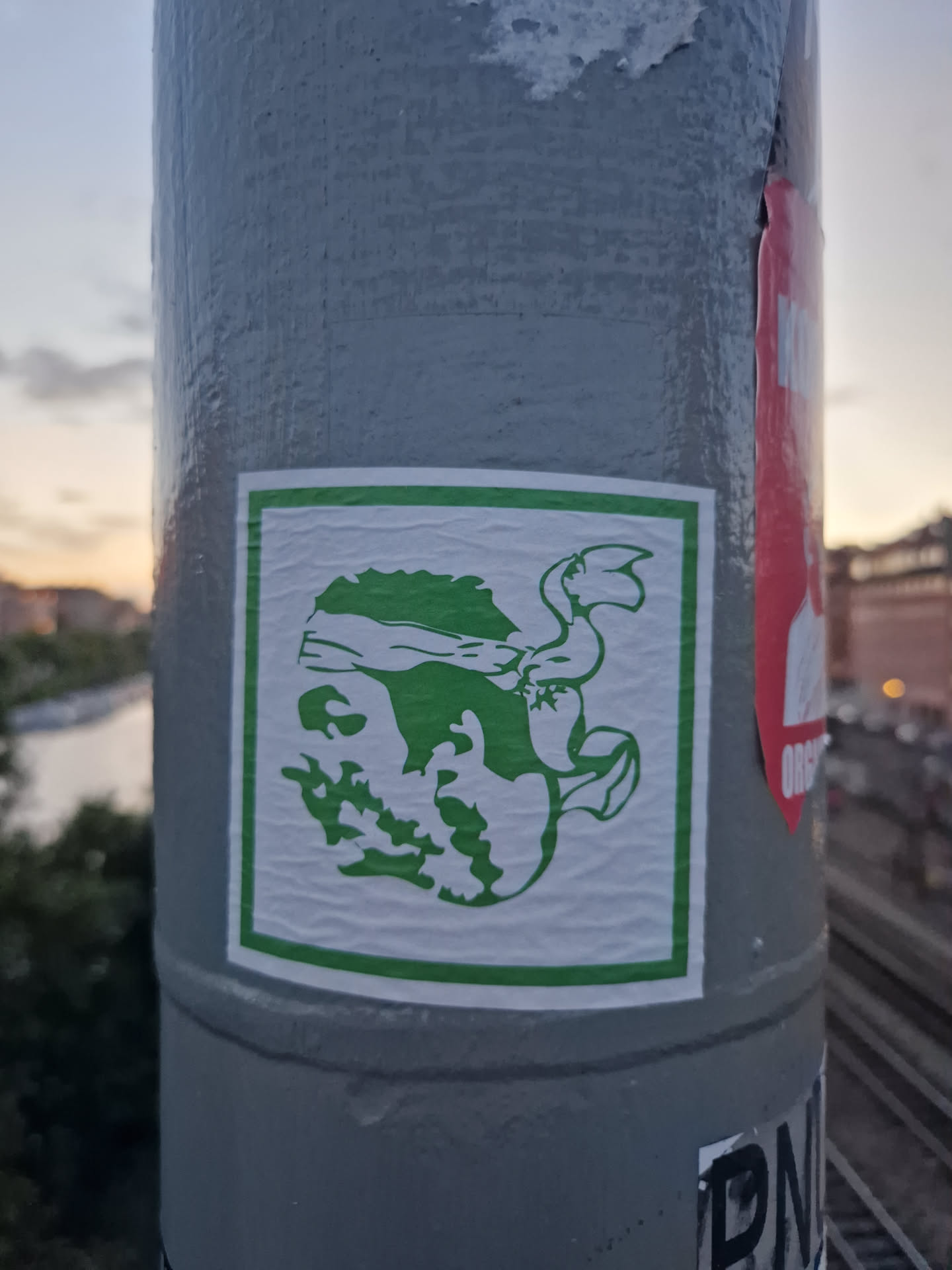 A green creature is depicted in a minimalist style on a white background, framed by a green square. The creature appears to be a stylized figure with a headband or bandana and possibly wings or appendages. The style suggests a possible reference to street art or graphic design. The sticker is affixed to a gray pole, likely outdoors.
