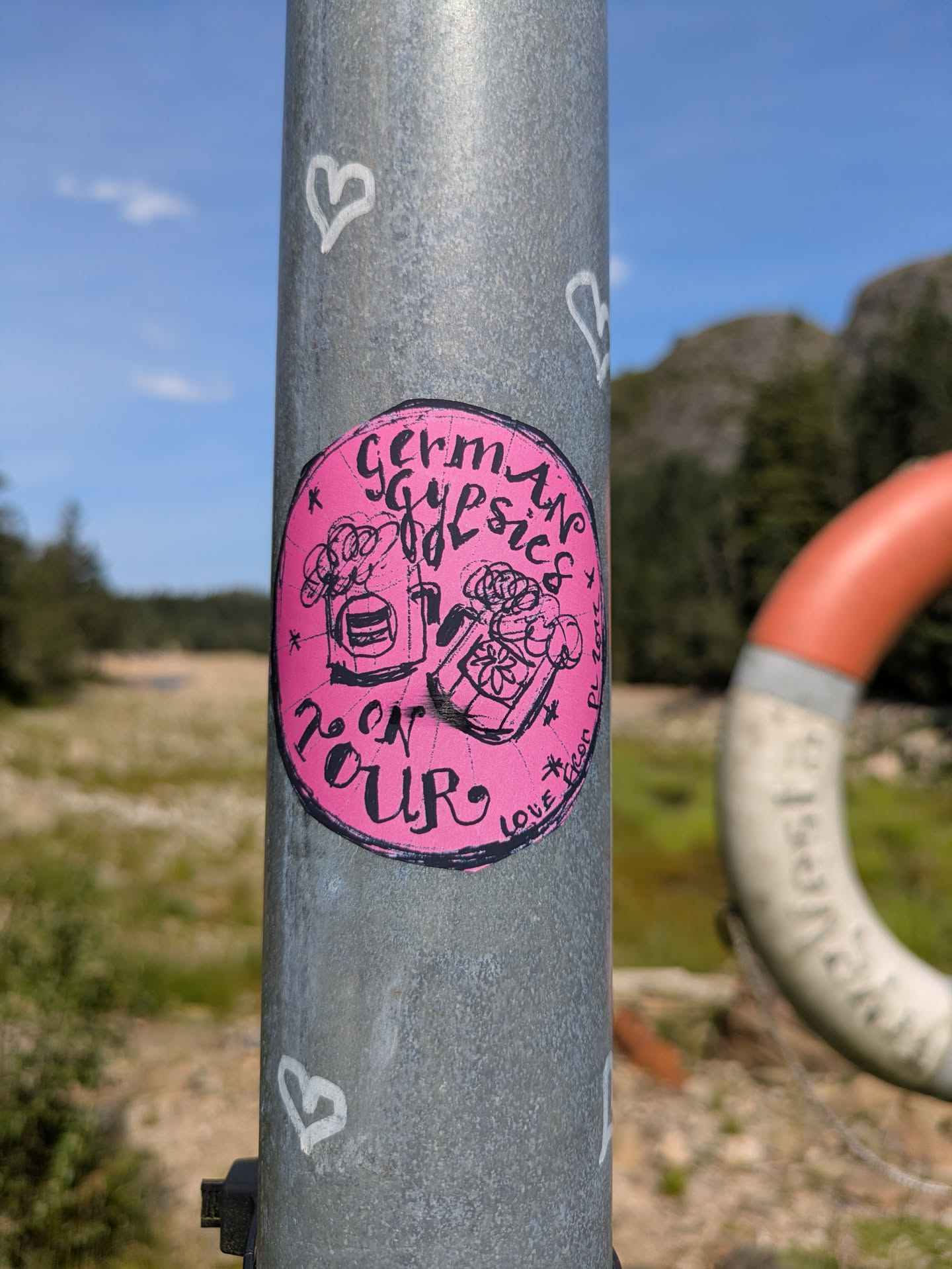 A circular, bright pink sticker affixed to a grey metal pole. The sticker features black, hand-drawn illustrations and text. On the left side, there's an image resembling a glass of frothy beer or a bubbly drink. On the right, there's a box or pot filled with flowers or plants. The prominent text at the top reads "German Gypsies" and at the bottom, "ON TOUR". Small decorative asterisks and hearts are scattered around the main artwork. Along the right edge, faint text reads "LOVE" and what appears to be a social media handle "@EICON_PL" followed by a partial date, possibly "202?". The metal pole also has white, hand-drawn heart shapes. The background shows a natural landscape with green fields, distant hills, a blue sky, and a blurred orange and white lifebuoy.