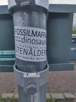 A sticker on a metal pole with text in Swedish. The text translates to: "Fossil Mafia consists of dinosaurs. They send us all to the Stone Age. Stop snorting their crap."