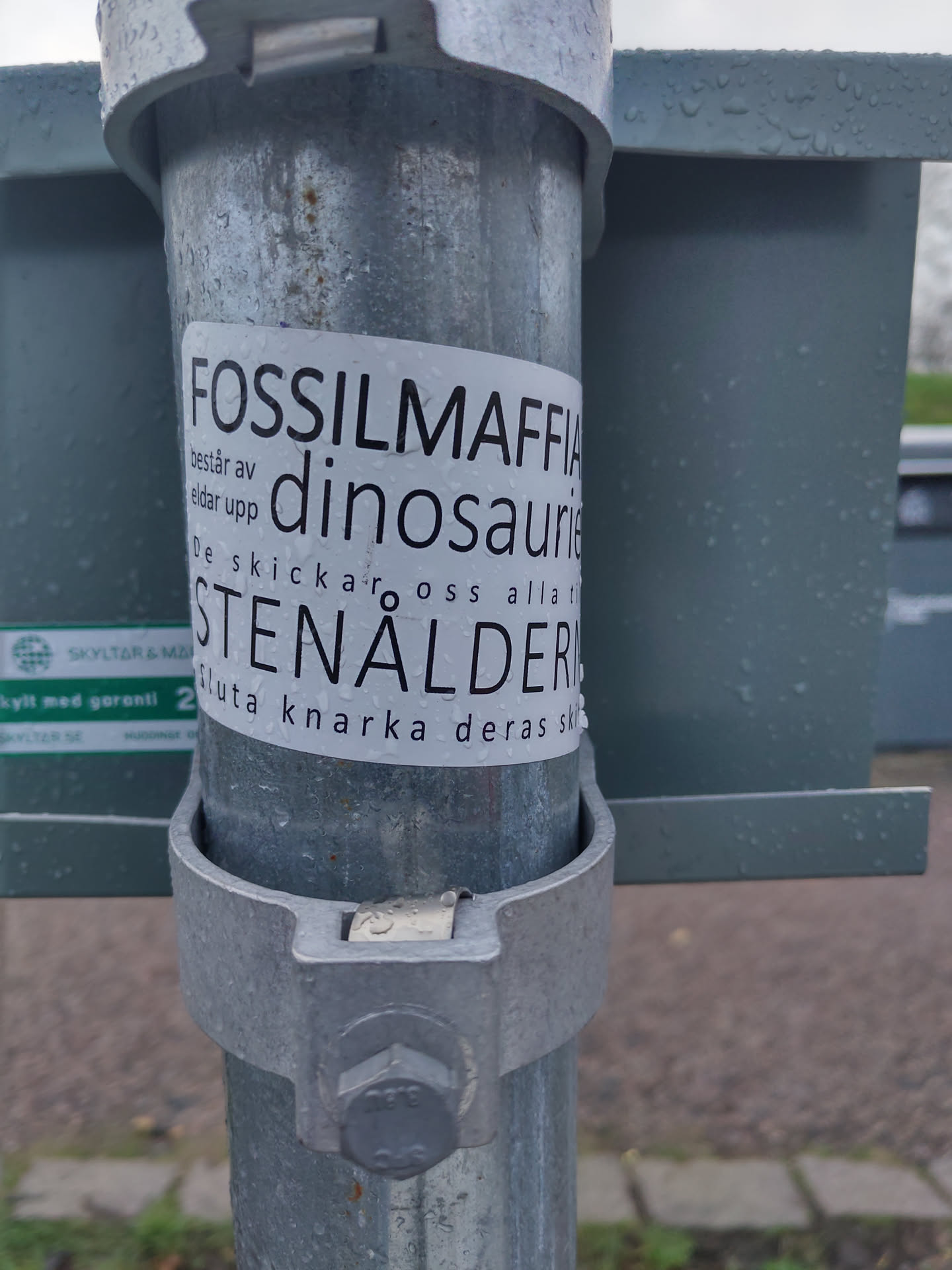 A sticker on a metal pole with text in Swedish. The text translates to: "Fossil Mafia consists of dinosaurs. They send us all to the Stone Age. Stop snorting their crap."