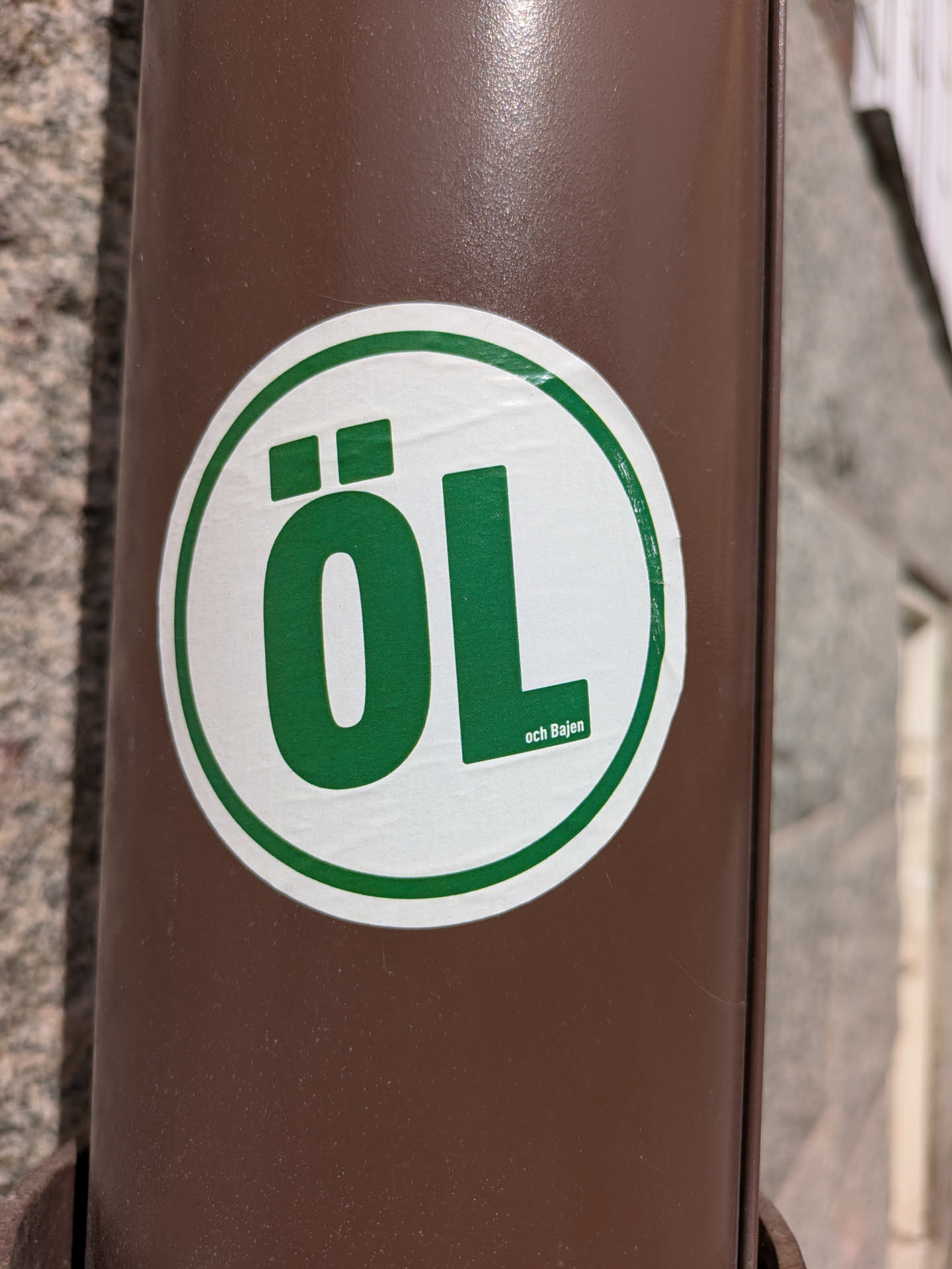 The sticker is a white circle with a green border, displaying the word "ÖL" in large, green letters. Below "ÖL", in smaller green text, it says "och Bajen". The sticker is affixed to a brown metal drainpipe.