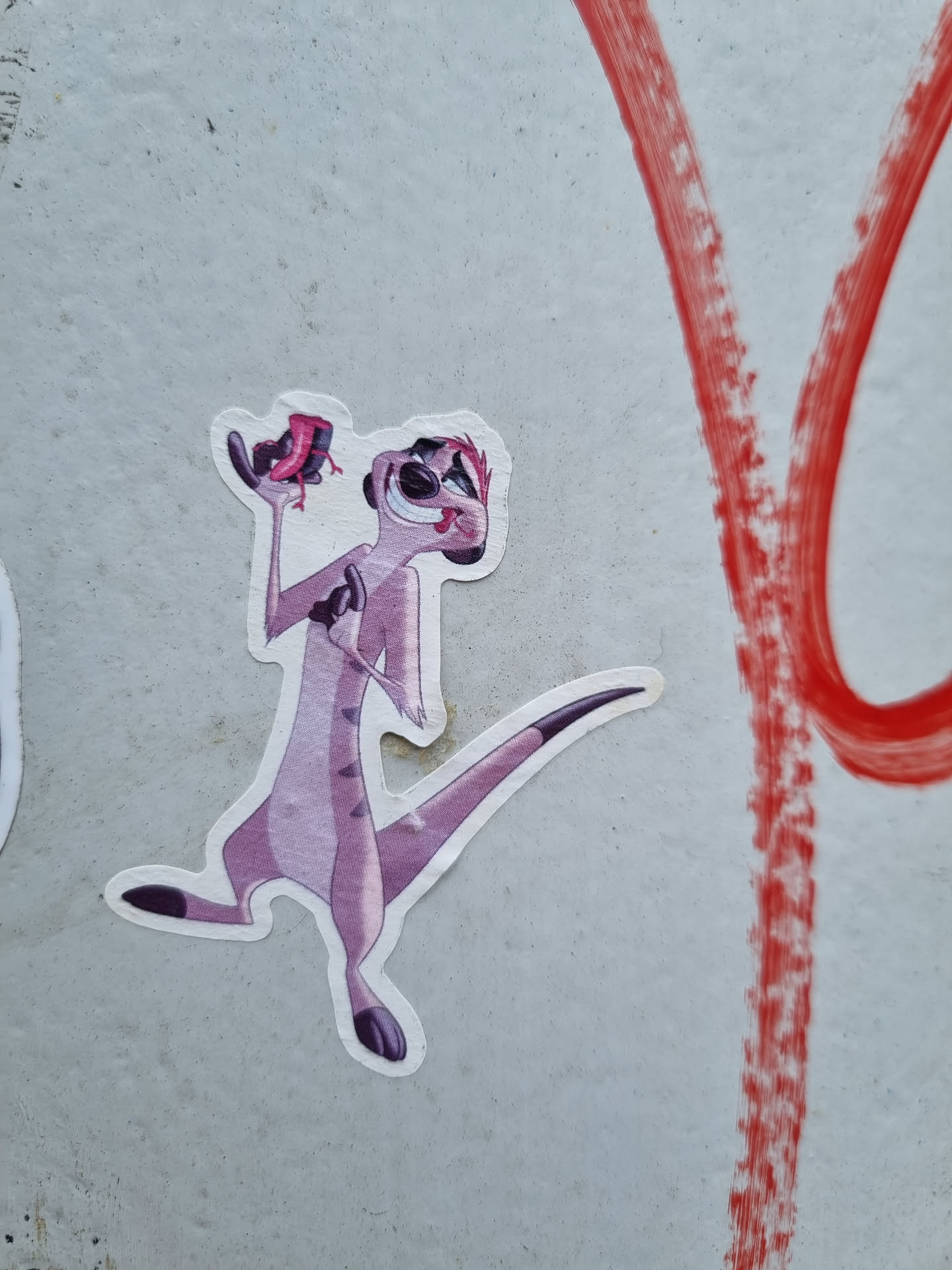 A sticker featuring Timon, the meerkat from Disney's The Lion King, holding what appears to be a large bug.  The sticker is placed on a wall with some red spray paint markings nearby.