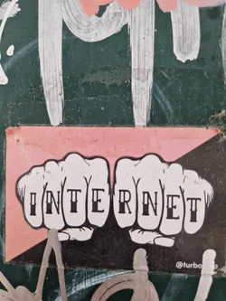 A sticker with two fists, each knuckle with a letter spelling out the word INTERNET. The sticker has a pink background and black and white illustration.  It's been stuck on a dark green wall with other graffiti.