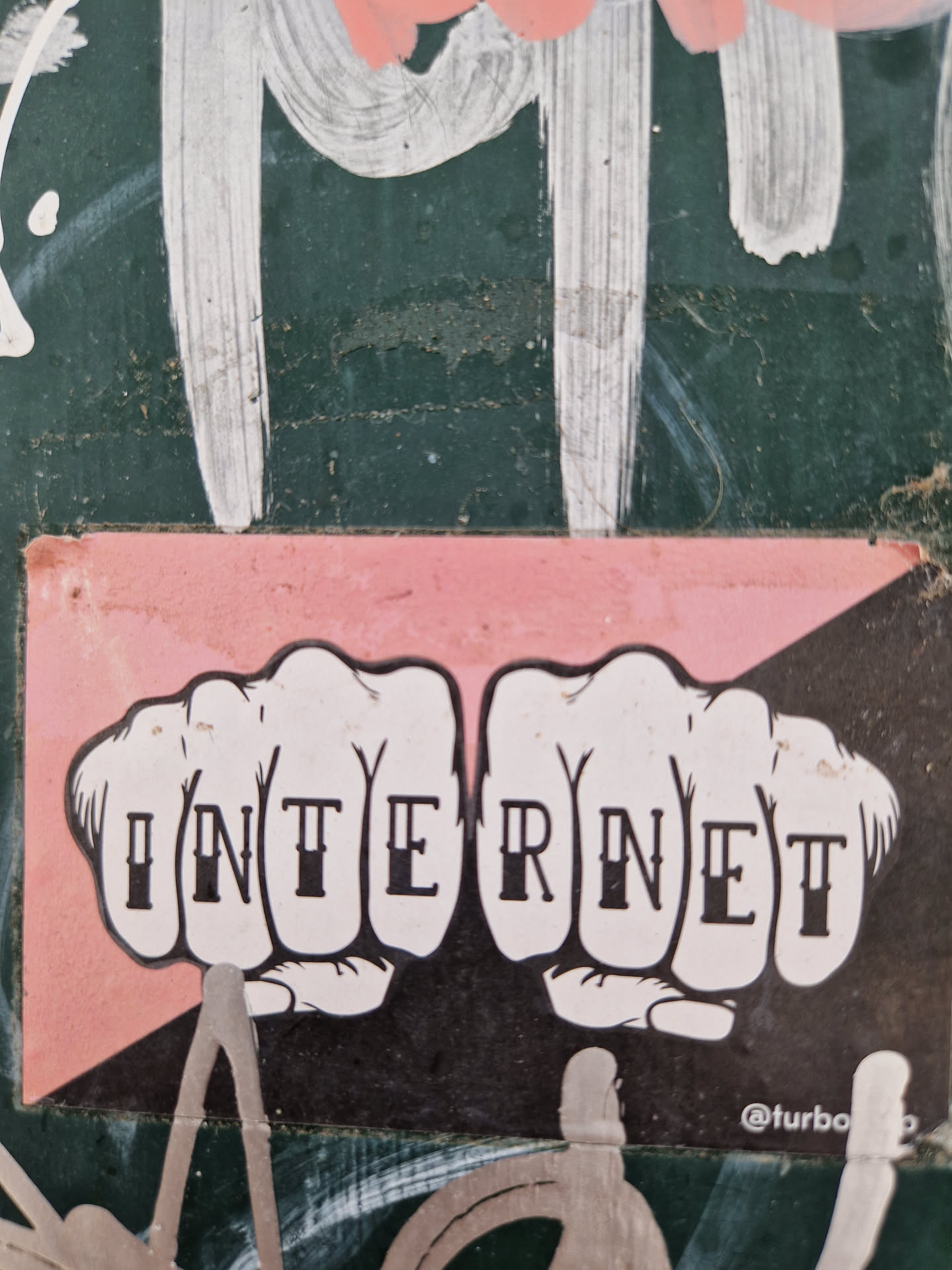 A sticker with two fists, each knuckle with a letter spelling out the word INTERNET. The sticker has a pink background and black and white illustration.  It's been stuck on a dark green wall with other graffiti.