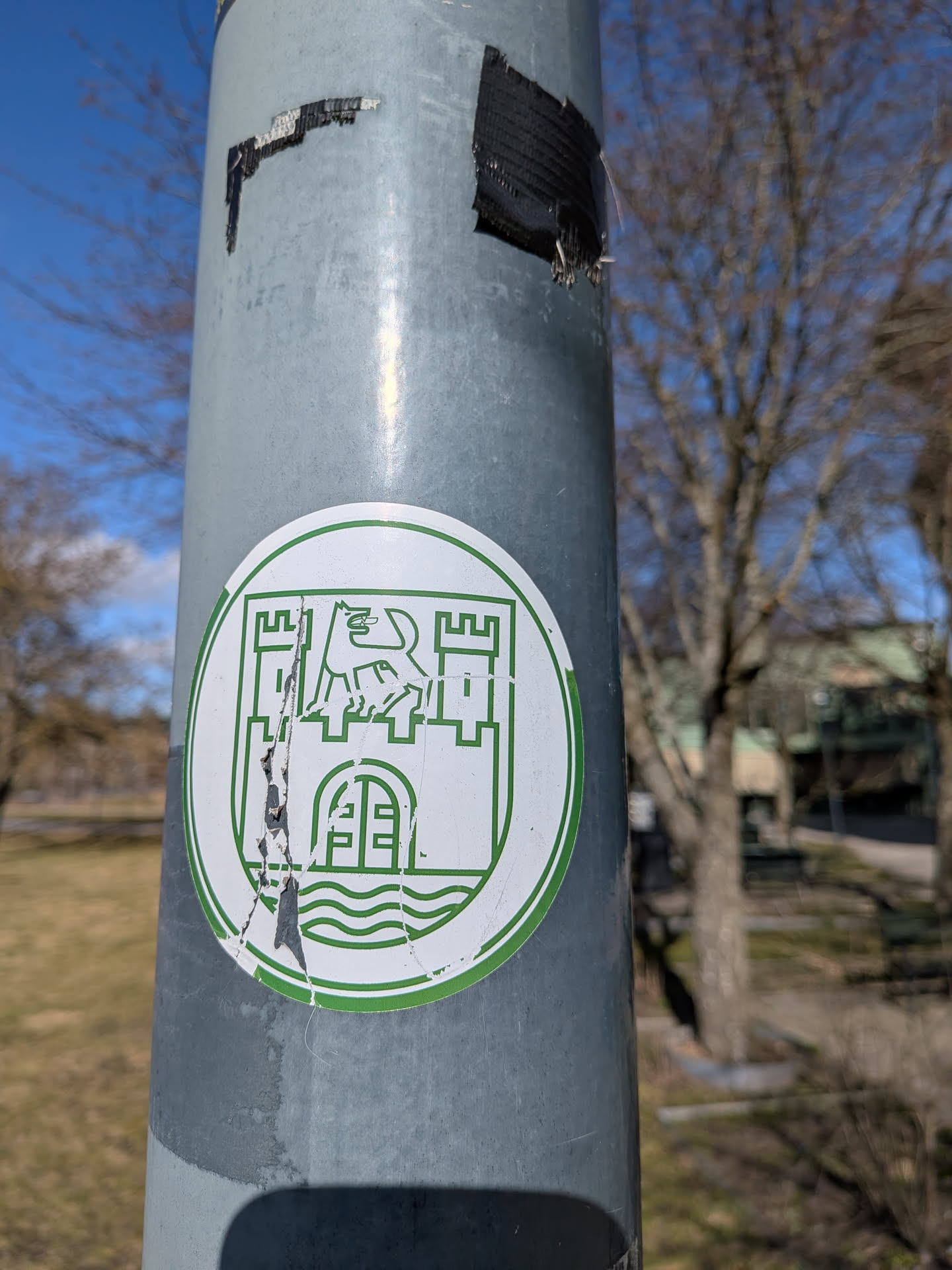 A circular sticker with a white background and a bold green outline. The central design is a stylized heraldic emblem. At the top, a rampant lion or griffin-like creature stands between two castle towers with battlements. Below this, within the lower part of the castle structure, is a gate or archway. The very bottom of the emblem features three wavy lines, suggesting water. The sticker shows signs of wear, including a prominent vertical tear through the left side of the design and some peeling around the edges.