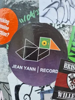 Round sticker with a geometric design resembling a bird, predominantly in black, white, and orange against a dark purple background.  The text 'JEAN YANN | RECORD' is visible at the bottom.