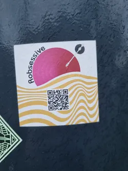 A sticker with a pink sun-like graphic, wavy lines, and a QR code. The word "Robsessive" is written vertically alongside the pink circle.