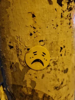 A small, round, yellow sticker depicting a sad face is affixed to a textured, weathered surface. The sticker's background is yellow, and the sad face is rendered in a simple style with black lines indicating the eyes, mouth, and tears.
