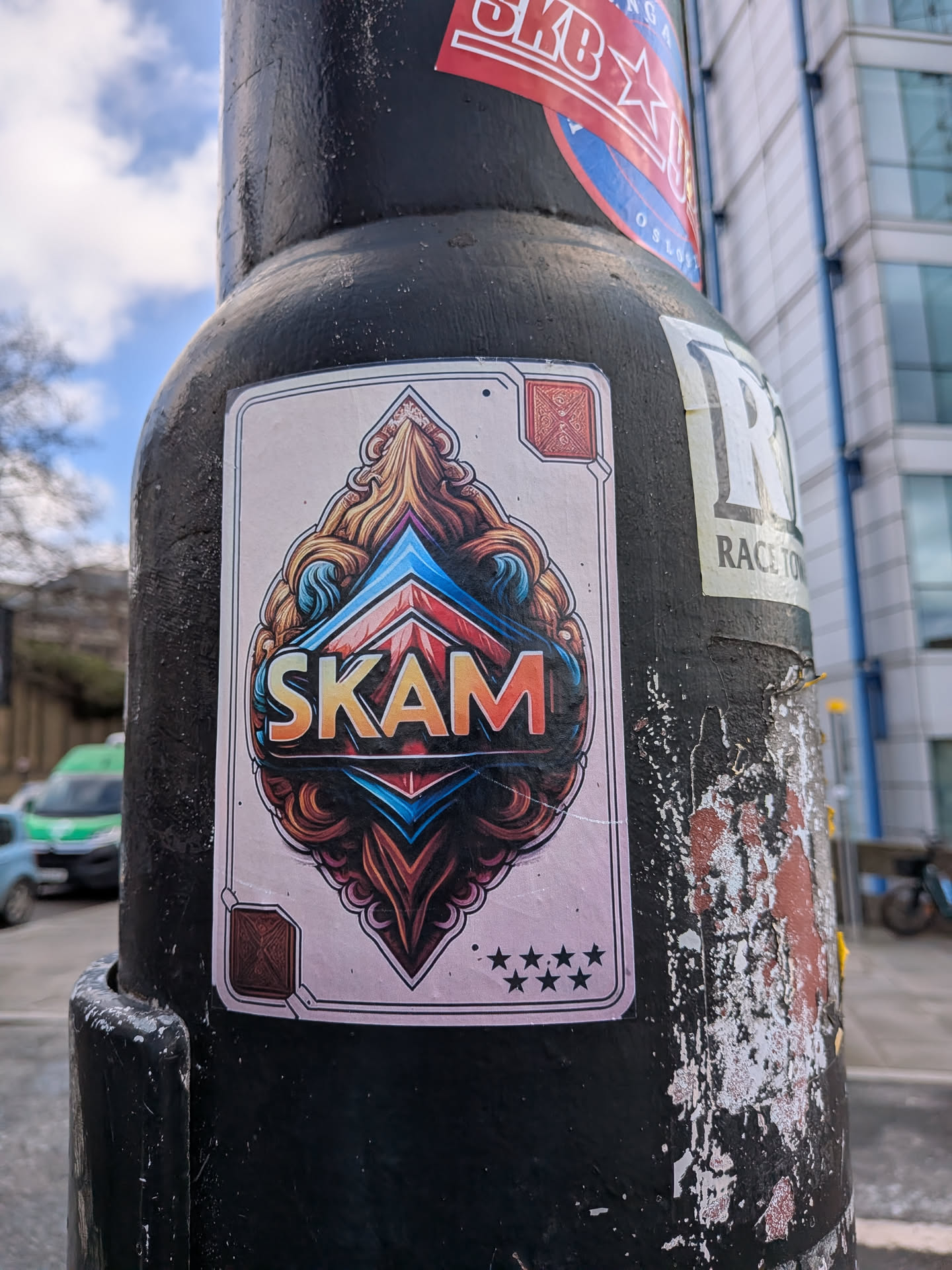 The sticker is rectangular with slightly rounded corners, designed to resemble an oversized playing card. It features a light, faded pinkish-beige background framed by a thin black double border. In the top and bottom corners, there are small, square, ornate symbols in a reddish-brown hue, reminiscent of stylized card suits. The central design is highly intricate and symmetrical, featuring a large, organic, almost flame-like or swirling pattern in shades of gold, brown, and orange. Within this ornate frame, a prominent, angular, diamond-like structure in vibrant blue and red provides a sharp contrast. The word "SKAM" is boldly displayed in the center, rendered with a gradient from orange to yellow, outlined in black, and appearing to have a slight 3D effect. Below the main design, near the bottom border, five small black stars are arranged horizontally. The overall aesthetic combines detailed graphic design with a street art sensibility.
