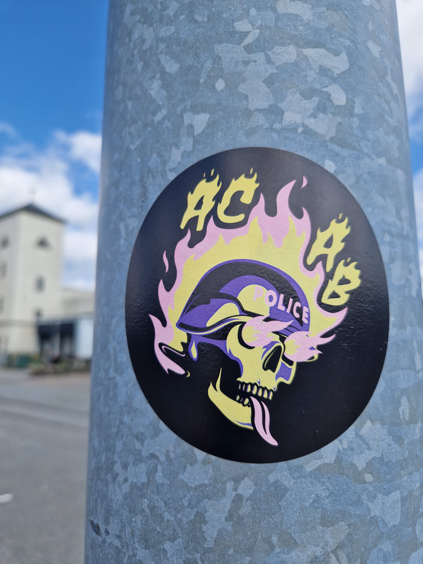 A round sticker with a skull wearing a police helmet engulfed in flames. The acronym ACAB is prominently displayed above the skull. The sticker is affixed to a metal pole.