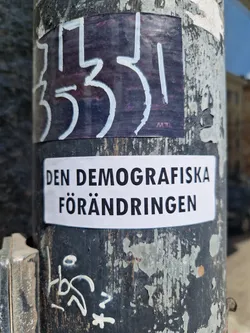 A white sticker with black text on a weathered pole. The text on the sticker translates to "The Demographic Change" in English. There's another sticker on top of it with illegible markings.