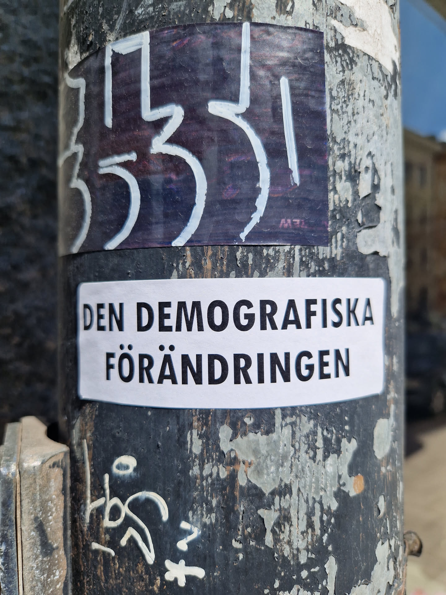 A white sticker with black text on a weathered pole. The text on the sticker translates to "The Demographic Change" in English. There's another sticker on top of it with illegible markings.
