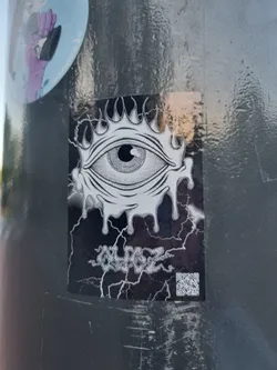 Black and white sticker with a detailed drawing of an eye surrounded by flames, dripping with a melting effect.  The sticker has a distressed or cracked background. Text below the eye appears to say "ZGLPGZ". A QR code is visible in the bottom right corner.