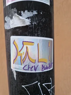 A holographic sticker with abstract gold and purple characters.  The artist's signature, "Chew Bebt," is visible in purple. The sticker features an iridescent finish. It's adhered to a dark-colored pole, possibly metal.