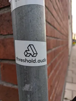 A white sticker with a geometric logo and the text 'threshold.audio' is affixed to a metal pole against a brick wall. The logo appears to be a stylized three-dimensional representation of the letter 'A'.
