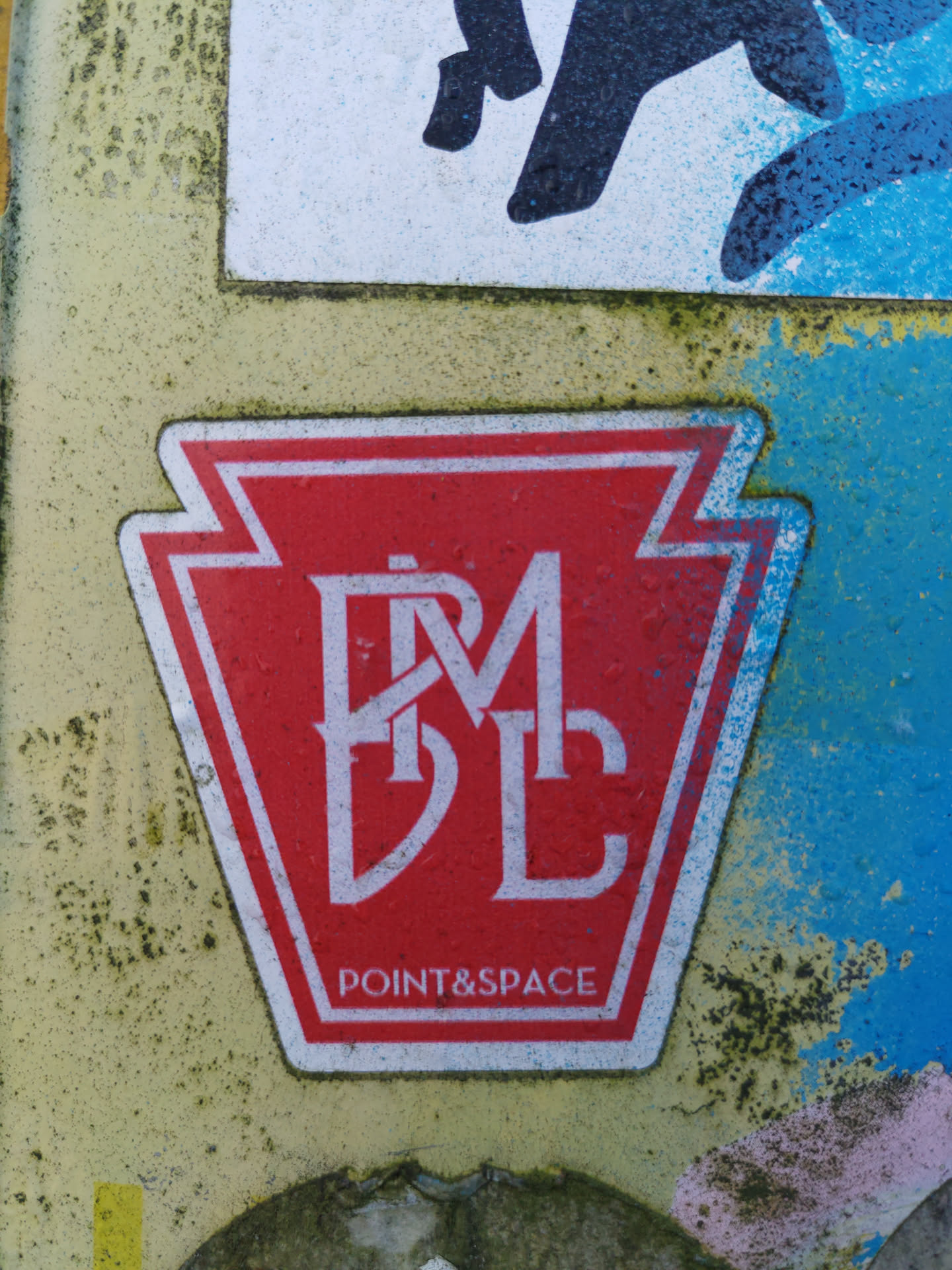 A red sticker in the shape of a pentagon with white trim. Inside, the letters P M D L are displayed in an elegant serif font. At the bottom, the text 'POINT&SPACE' is written in a smaller font. The sticker is affixed to a weathered surface with other stickers and paint visible around it.