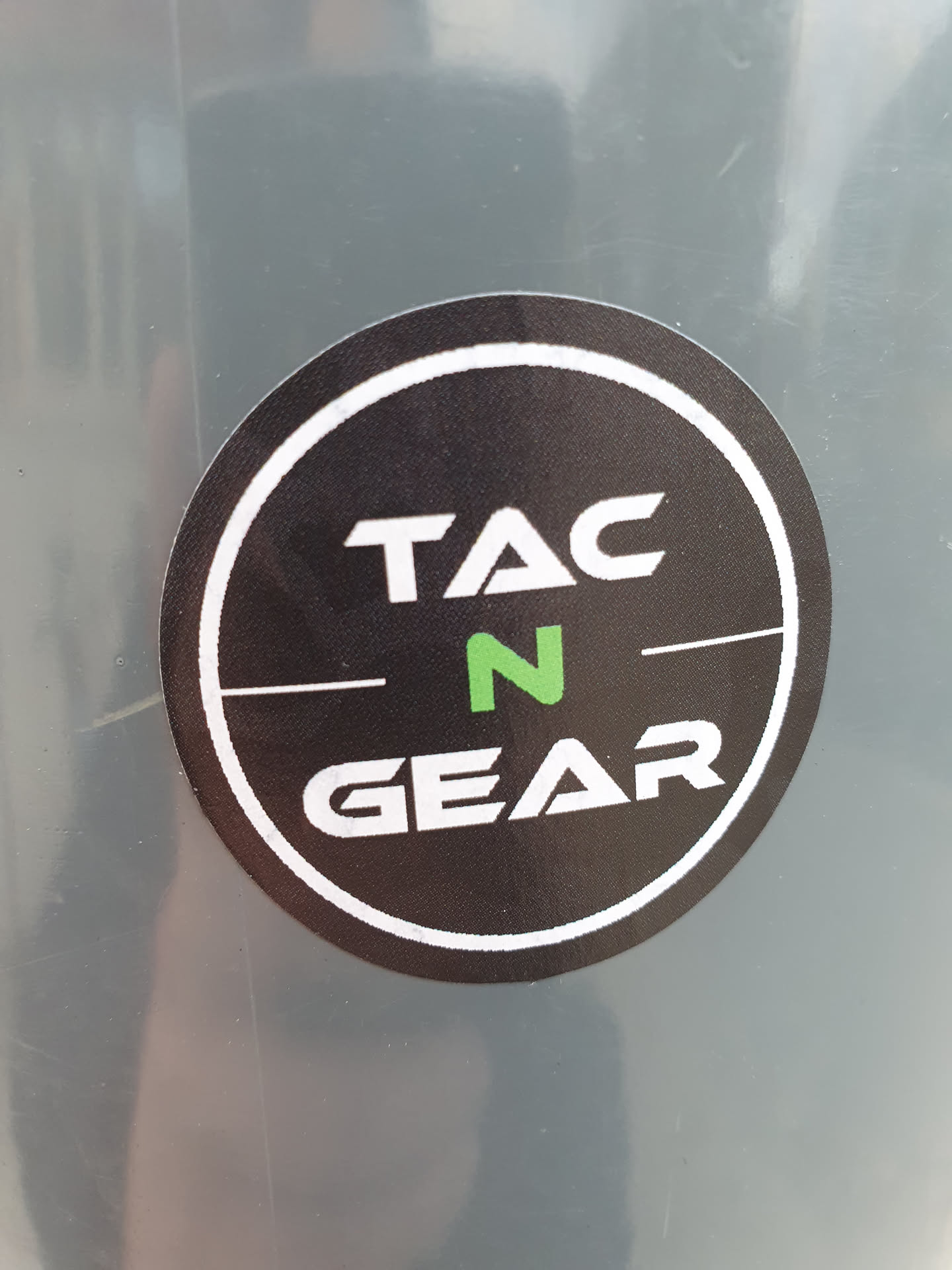 A round sticker with Tac N Gear written in a circle. The text is in white with a green N. The background is black, and it has a white border.