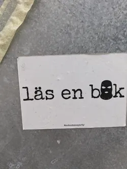 A minimalistic sticker with the text "Läs en bok" (Swedish for "Read a book") and a balaclava icon.  The sticker has a simple design on a white background.  The text is in a typewriter-style font.