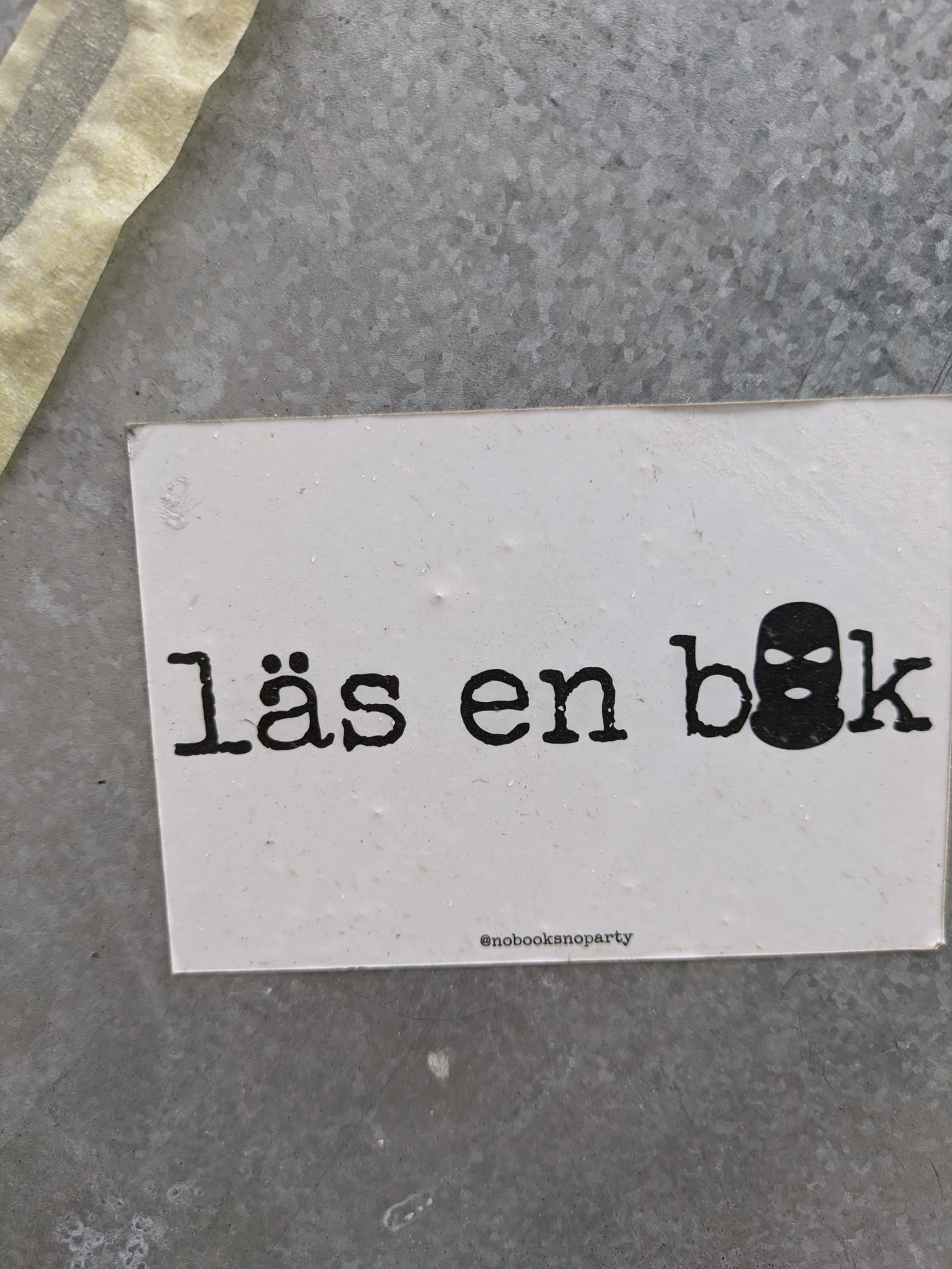 A minimalistic sticker with the text "Läs en bok" (Swedish for "Read a book") and a balaclava icon.  The sticker has a simple design on a white background.  The text is in a typewriter-style font.