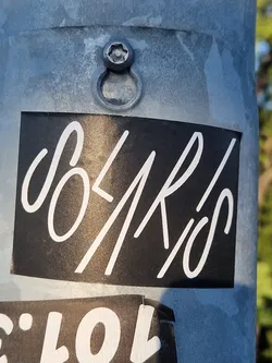 The image features a rectangular black sticker adhered to a textured, light gray metal pole. The sticker displays the word 'SOLARIS' in a flowing, elegant white script font, which contrasts sharply against the dark background. The font gives the word a distinctive, almost calligraphic appearance. Above the sticker, part of a metal fixture, possibly a bolt or hook, is visible on the pole. Below the main sticker, the top edge of another partially visible sticker with white text is present.