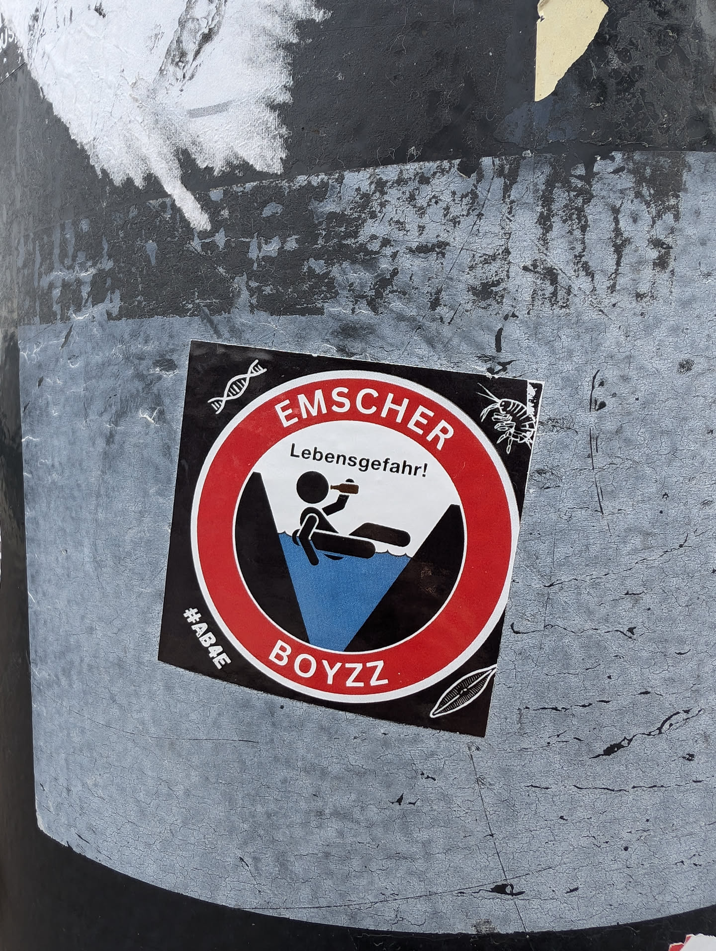 A round sticker with a red border and a black center. It has a pictogram of a person falling into water and the text EMSCHER Lebensgefahr! (EMSCHER Danger of death!). There is also the text #ABAE BOYZZ at the bottom. The sticker is affixed to a dark-colored surface.