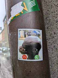 A rectangular sticker simulating a smartphone's incoming call screen. At the top, in a white box, the text 'Bruh Is calling...' is displayed in black. Below this, a large, somewhat blurry image of a person's dark-skinned, bald head is shown in profile, set against a blurry background of buildings. At the bottom left, there's a small alarm clock icon with 'Remind Me' text, and at the bottom right, a small white circle with 'Message' text. Below these options are two prominent circular buttons: a red one on the left with a white phone hang-up icon, and a green one on the right with a white phone answer icon. The sticker has a white border and appears slightly worn.