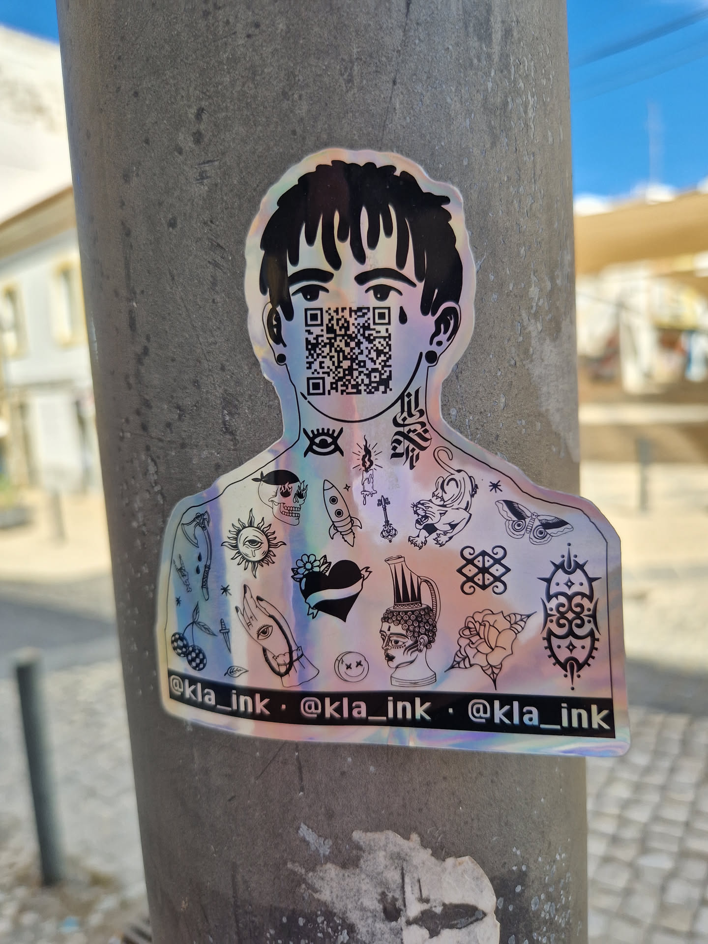 This iridescent sticker showcases a stylized character adorned with a collection of tattoos across their chest and shoulders.  Their face is obscured by a QR code. The sticker has a holographic or iridescent finish. The bottom of the sticker includes the Instagram handle @kla_ink repeated three times.