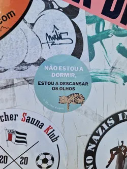 A cute sticker with a sleeping cat and a Portuguese phrase that says: "I'm not sleeping, I'm resting my eyes." It's a light teal circle with a simple illustration. The phrase is written in a sans-serif font. The small text at the bottom says "o teu mal é sono", which translates to "your evil is sleep".
