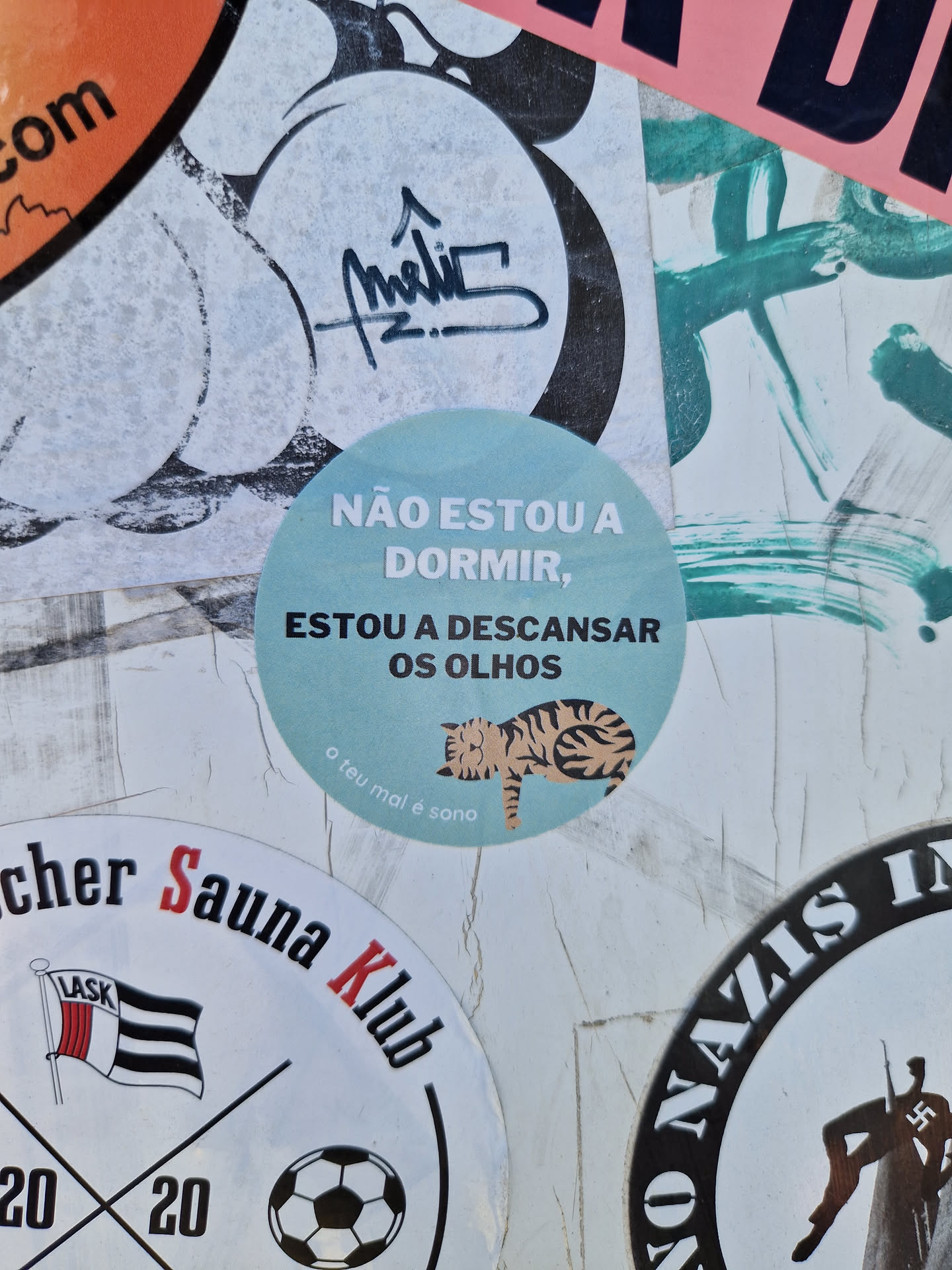 A cute sticker with a sleeping cat and a Portuguese phrase that says: "I'm not sleeping, I'm resting my eyes." It's a light teal circle with a simple illustration. The phrase is written in a sans-serif font. The small text at the bottom says "o teu mal é sono", which translates to "your evil is sleep".
