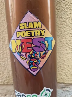A colorful diamond-shaped sticker with the text "Slam Poetry Nest" and a stylized illustration of a mask or face. The text is in a bold font with each letter in a different color. The illustration is colorful and intricate.