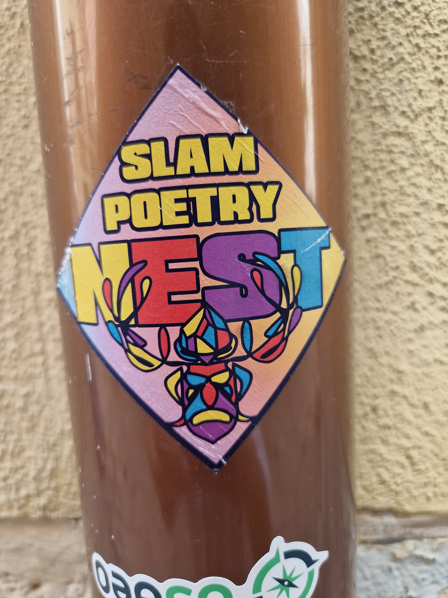 A colorful diamond-shaped sticker with the text "Slam Poetry Nest" and a stylized illustration of a mask or face. The text is in a bold font with each letter in a different color. The illustration is colorful and intricate.
