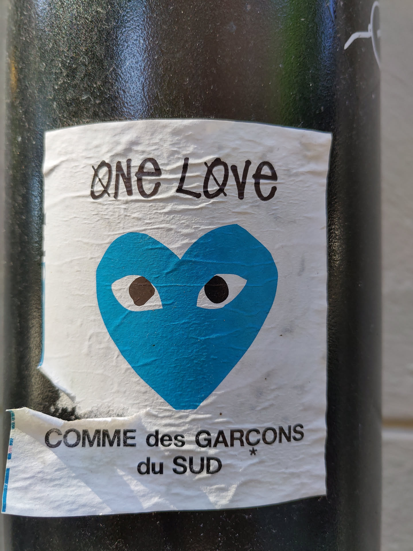 A sticker featuring a blue heart with eyes and the text 'One Love' above and 'Comme des Garçons du Sud' below. The sticker appears to be affixed to a dark-colored pole.