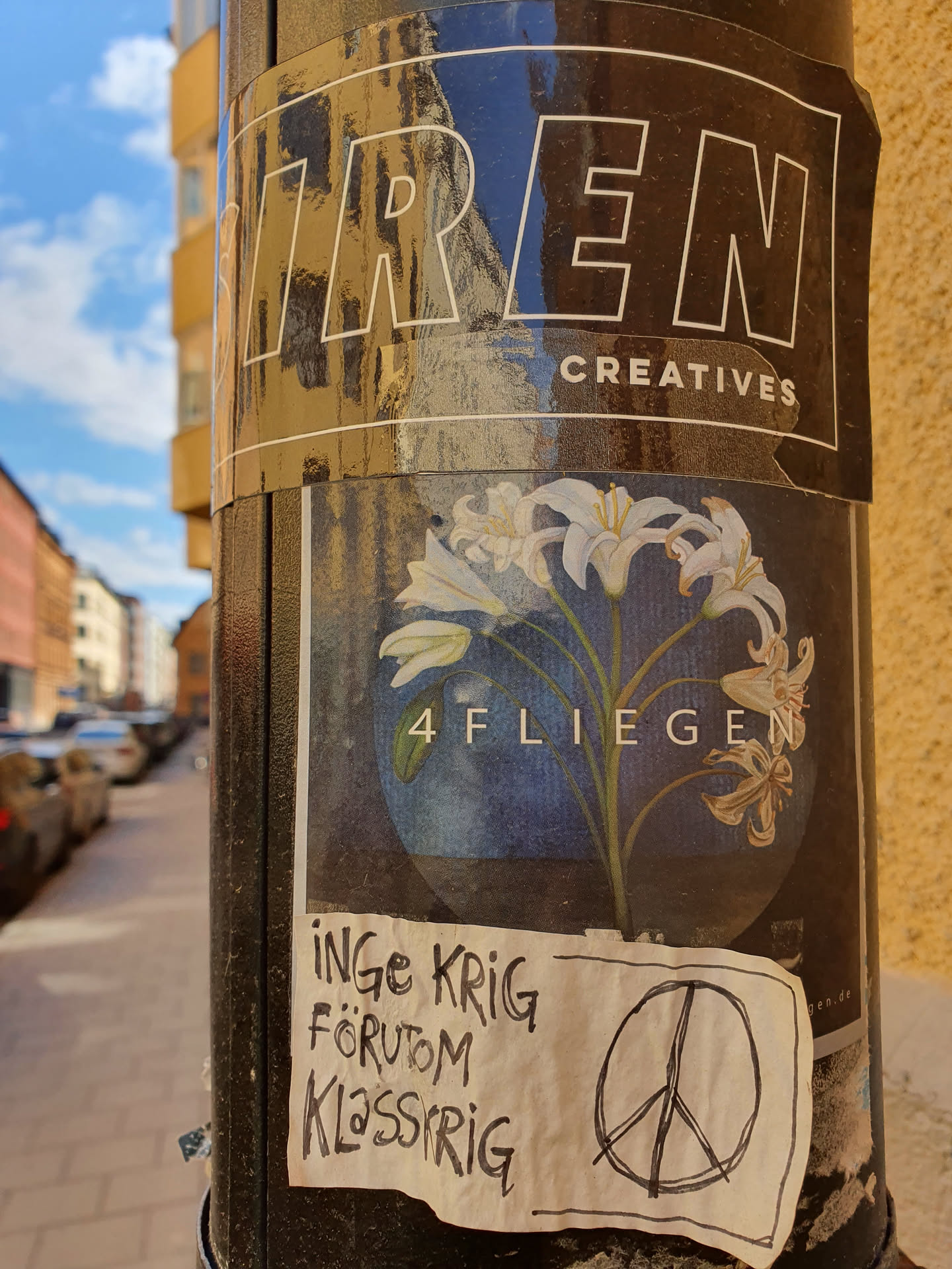 A small, hand-drawn sticker featuring a peace symbol and the text  "Inge krig förutom klassrig" (Swedish for "No war except class war") is placed on a pole, which also has other stickers. Among them, a sticker with an illustration of white lilies and the number 4FLIEGEN is visible. 