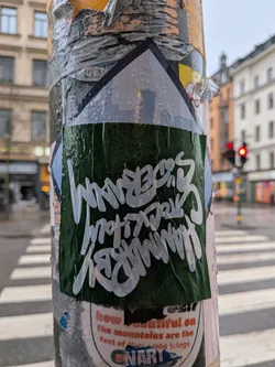 The sticker is rectangular with a pointed top, resembling a house or arrow shape, and is affixed to a metal pole covered in other worn stickers and tape. The main body of the sticker is a dark, forest green color, appearing somewhat textured and wet with numerous water droplets. Overlaid on this green background is stylized white text, written in a distinct graffiti handstyle. The text reads 'WANDERS' on the top line, 'STOCKHOLM' on the middle line, and 'GRAFFITI' on the bottom line. The white lettering has a slightly translucent quality, allowing some of the green background to show through, especially where the water droplets are. The sticker is slightly creased and shows signs of wear, indicating it has been exposed to the elements.