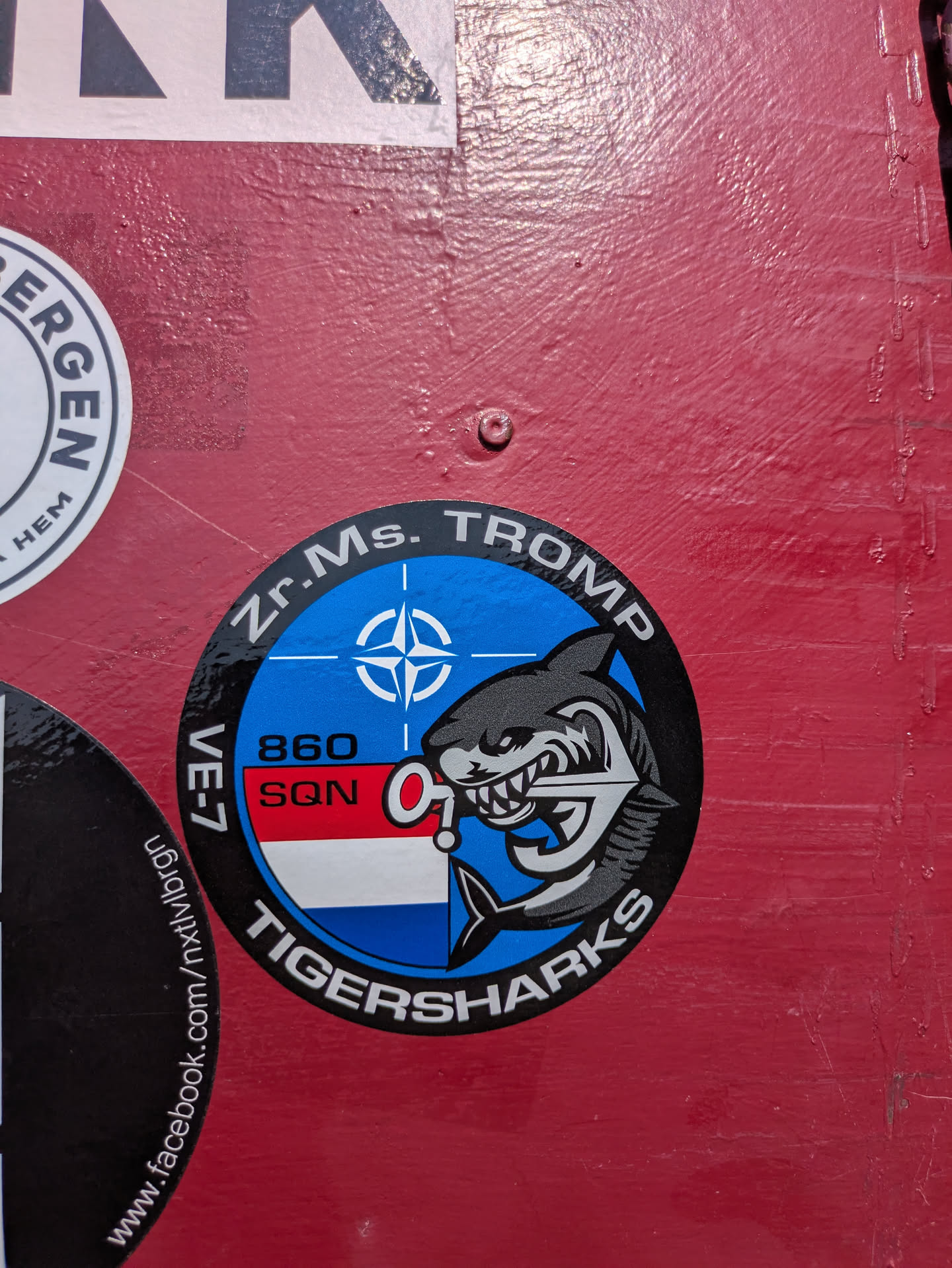 A circular sticker featuring the insignia of the Royal Netherlands Navy's 860 Squadron. The design is split into an upper blue section with a white NATO compass rose symbol, and a lower section with horizontal red, white, and blue stripes resembling the Dutch flag. A fierce grey tiger shark with bared teeth, holding an anchor, emerges from the right side. The outer black ring contains the text "Zr.Ms. TROMP" at the top and "TIGERSHARKS" at the bottom. "860 SQN" is written in the center-left, with "VE-7" partially visible on the left edge. The sticker likely represents a specific naval unit or ship.