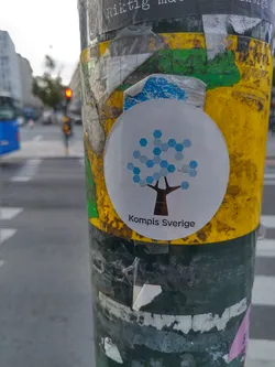 A round sticker featuring a stylized tree made of hexagons in shades of blue on a white background.  The text "Kompis Sverige" is printed beneath the graphic. The sticker is affixed to a weathered pole, surrounded by other old stickers.