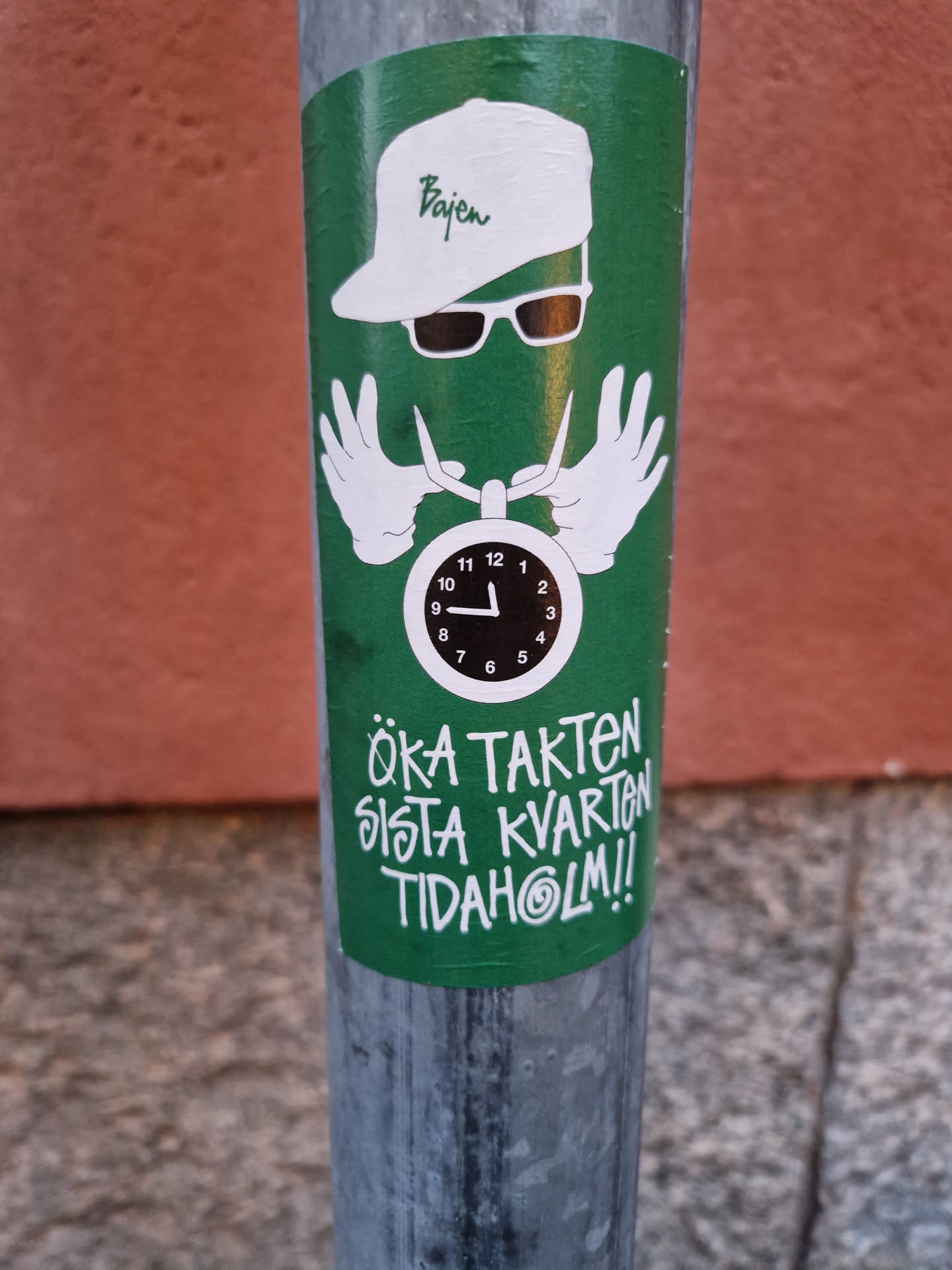 A green sticker featuring a unique design. The central figure has a "Bajen" branded cap and sunglasses, hands forming the shape of a clock's hands. The text below translates roughly to: "Increase the pace last quarter Tidaholm!!"
